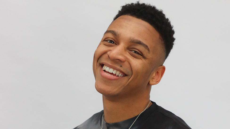 New Radio 1Xtra presenter DJ Day Day's headshot photo. He is smiling at the camera and has his head tilted slightly to the right. He has short black hair and is clean shaven. We can only see the tops of his shoulders but he has a thin silver necklace on and is wearing a black jumper.