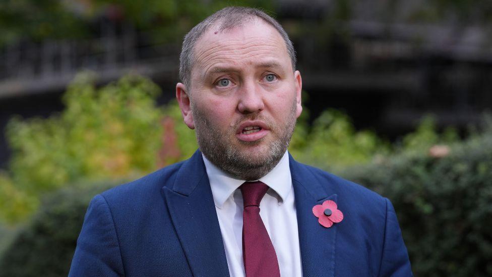 Ian Murray wearing a blue suit, red tie and red poppy while being interviewed, with trees and bushes in the background  