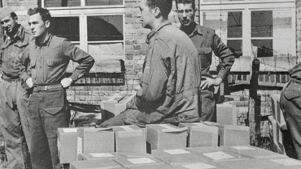 A black and white World War Two picture of prisoners of war next to small cardboard boxes with food in them