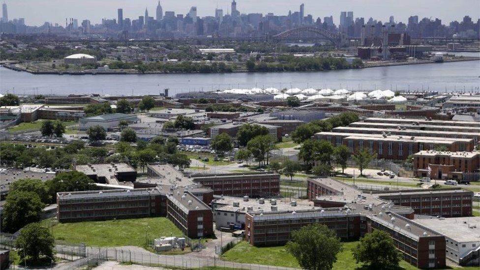 The Rikers Island jail complex stands in the foreground with the New York skyline in the background.