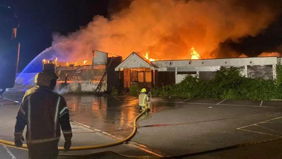 The club's clubhouse is well alight, with fire pouring from the single-storey building, with a firefighter spraying water from a hose close to the building, as another firefighter watches on