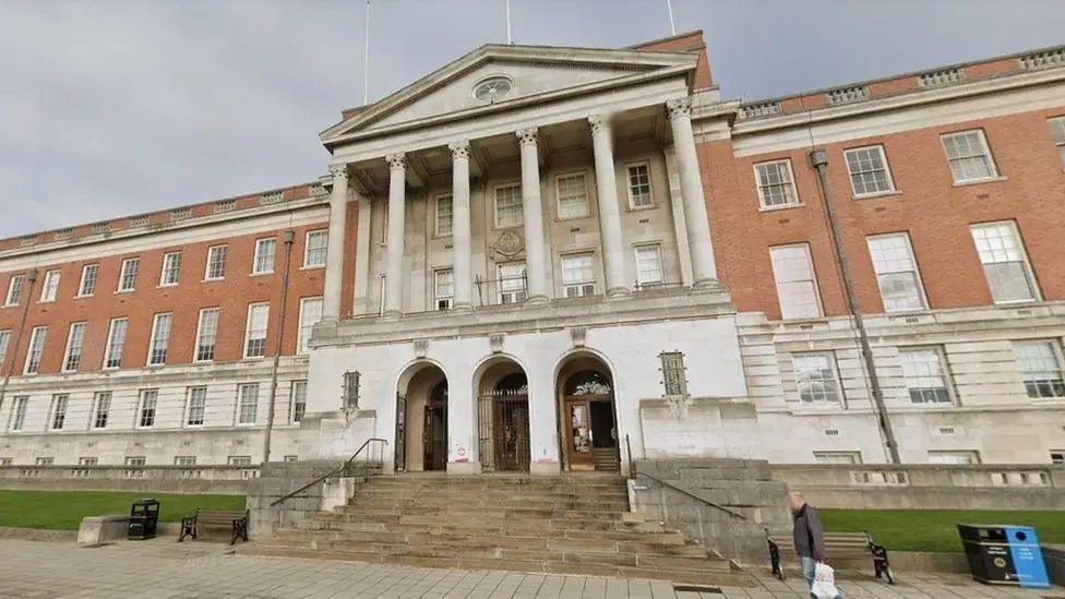 An imposing looking council office building in red and white stone with six columns above a triple doorway and a wide stairway from the pavement below