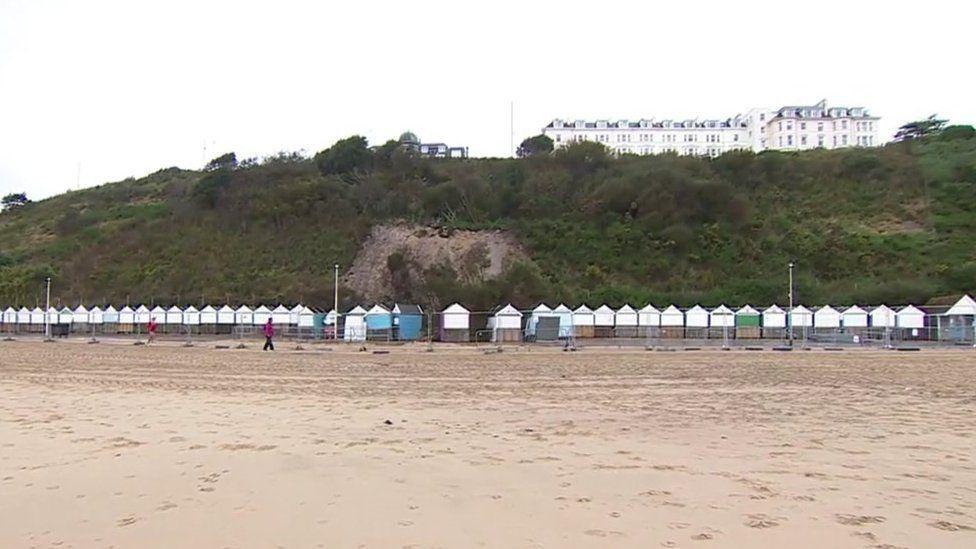 Golden sandy beach with footsteps line of white-topped beach huts, cliffs behind and a large white building on the cliff top.