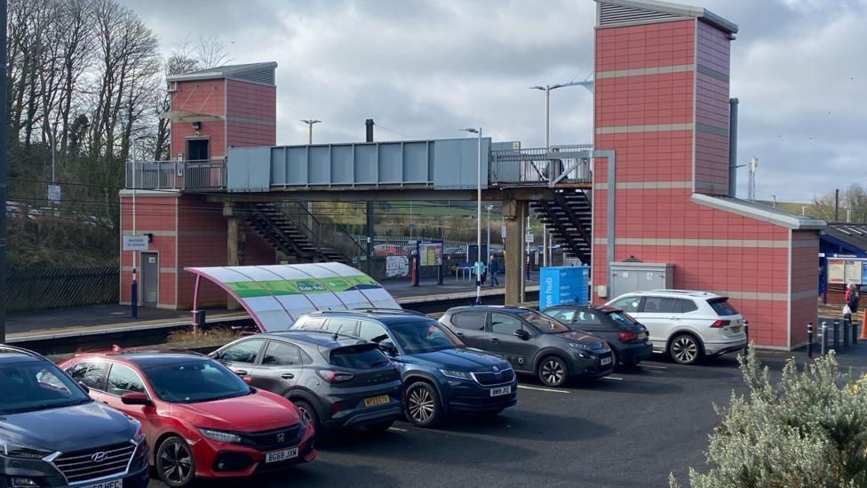 A picture of Alnmouth station with a number of cars parked up. Two pink towers contain a lift which links to a bridge over the  line 