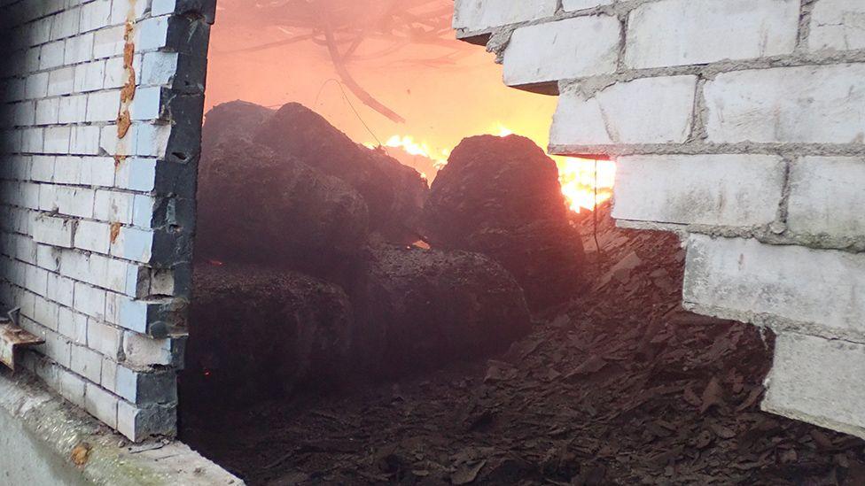 Waste on fire seen through a hole in a brick wall on the waste recycling site.