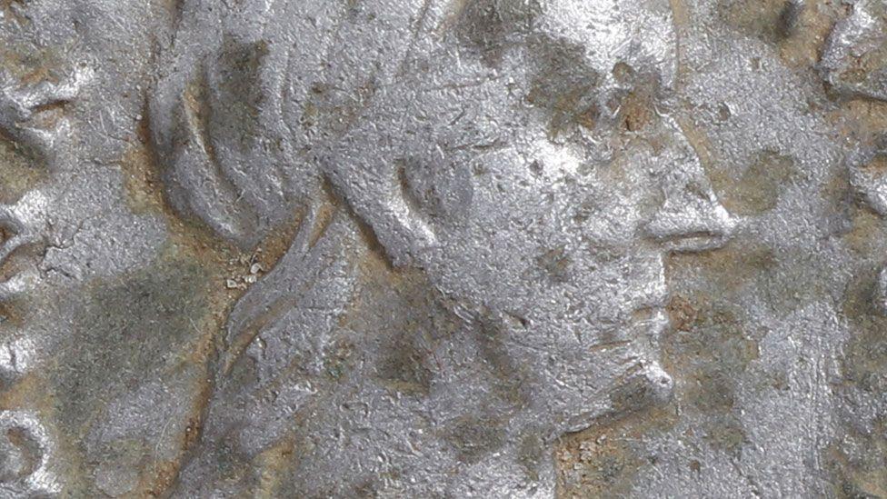 A close-up of a silver Roman denarius. It shows the empress Sabina in profile, facing right. She has a large slightly hooked nose. Her hair is pulled back behind her ears and locks flow down her neck. The coin is rather pitted. On the left can be glimpsed some of the letters which in full spell out her name.