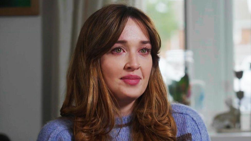 Kate Driver, with long, wavy auburn hair wearing a light purple knit sweater is seated indoors. The background shows a bright window with sheer curtains, some greenery outside, and a few decorative items on the windowsill, including a small plant and a candle holder.