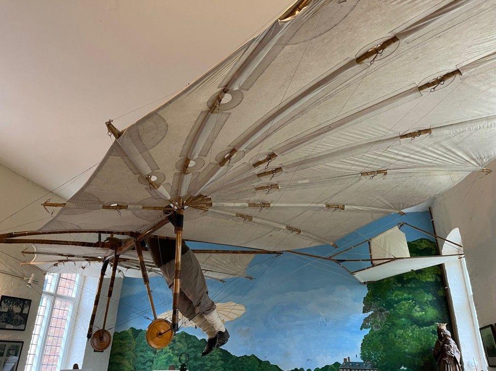 A replica of the Hawk glider is suspended from the ceiling at Stanford Hall. Behind it is colourful artwork depicting a clear blue sky and the tops of green trees.