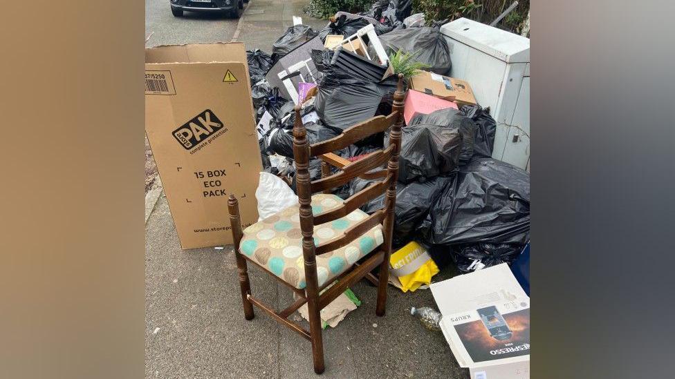 A pile of black bin bags on a pavement, with some split open and rubbish on the floor. There are cardboard boxes on the pavement, and a wooden chair as well.