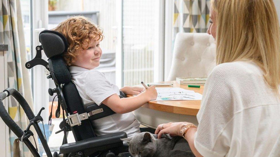 A smiling child in a wheelchair sitting at a desk and drawing beside a teacher