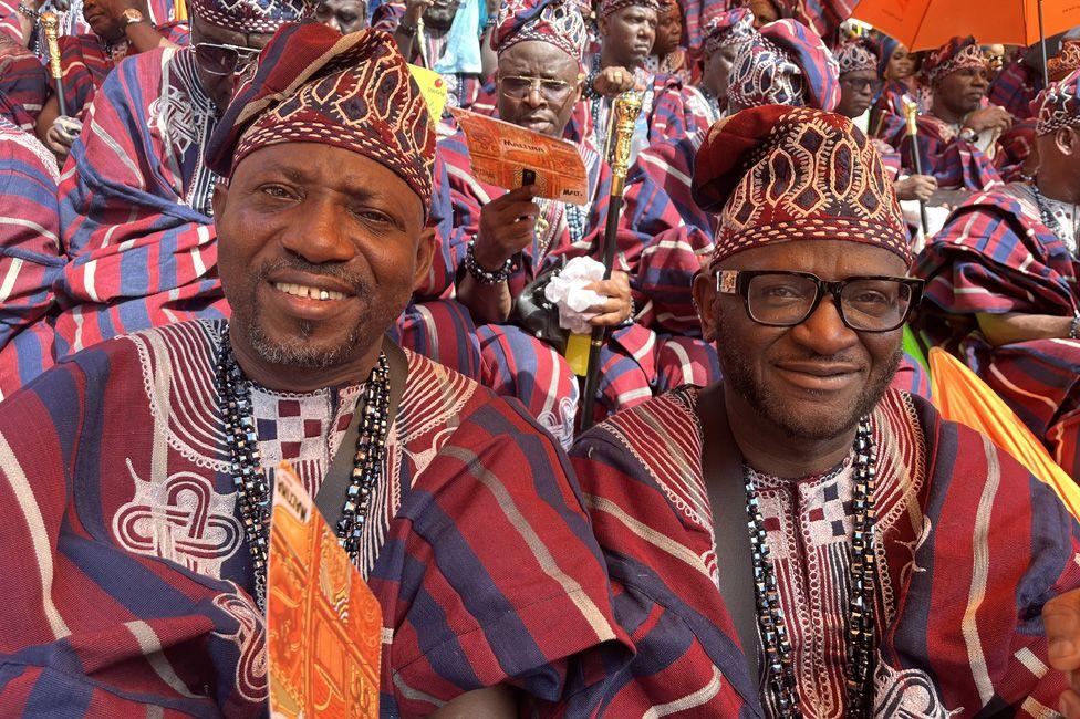 Two men in matching traditional outfits sitting in the stadium seats surrounded by others in similar clothes. 