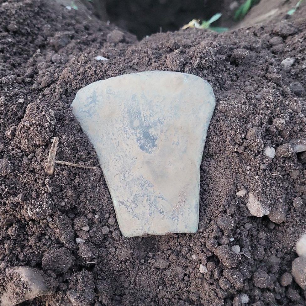 Old axe head covered in dust, sitting on dirt, in a field, next to a hole.