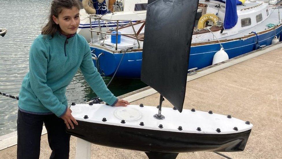 Anahita Laverack holding the boat, standing on a long slender black keel with a black flat sail, on a marina dock