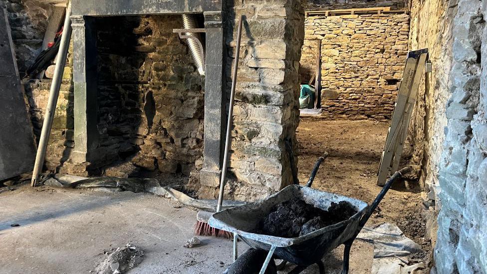 Stone walled interior with concrete and mud floors, a mantlepiece, broom, and wheelbarrow containing mud.