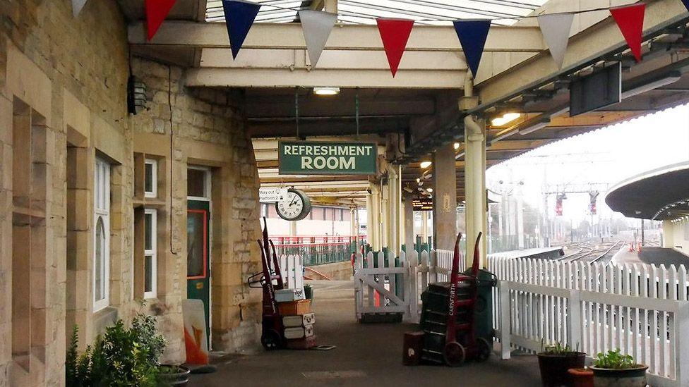 The platform at Carnforth station showing 1940s suitcases on trolleys, red white and blue bunting and a sign hanging over the platform saying Refreshment Room