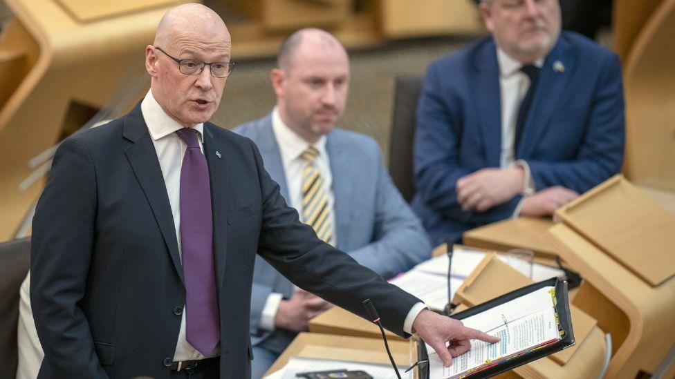 John Swinney, who is bald, speaking in the Scottish Parliament. He is wearing a dark suit, white shirt and purple tie. He is visible from the waist up and is pointing to a folder on the podium in front of him with his left hand. Two men are visible in the benches behind him. 