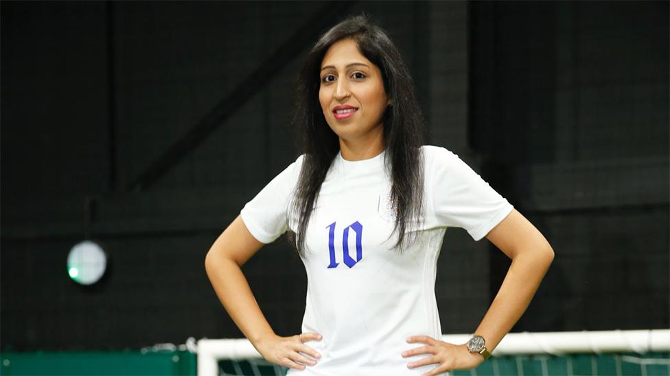 Sana, an Asian woman, wearing a white jersey with the number "10" written in blue in the middle. She has her hands on her hips, with a watch on her left wrist, and is looking at the camera with a smile. The background is dark.