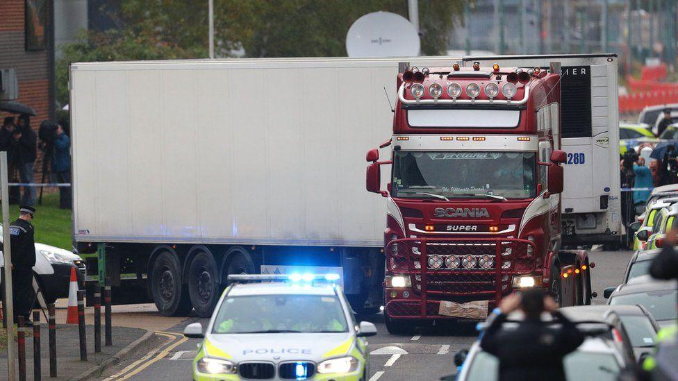 A lorry being escorted by a police car in front of it with traffic held up on the right. The lorry has a red cab and is pulling a white container, turning a corner with the body of the container at a right angle to the cab.