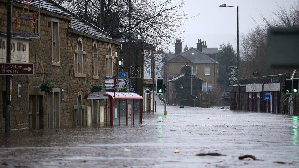 A street is submerged under flood water. There is a traffic light on green sticking out from the water in the centre of the picture, as well as the top half of a bus stop. Brick buildings on either side of the street are also submerged in water up to their bottom floor windows.