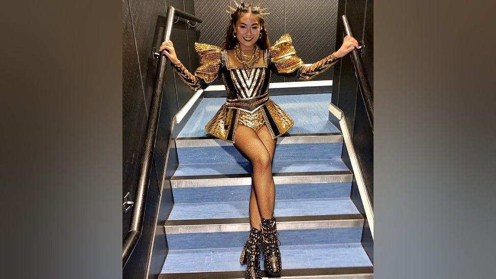 Jasmine is sitting on stairs , holding onto railings either side. She is wearing a spiked gold headdress. Her outfit is black, gold and sparkly. It has large padded shoulders with sparkles, and a flared mini skirt with a cut-out at the front, revealing a leotard underneath. Her boots are black, heeled with gold sparkles.