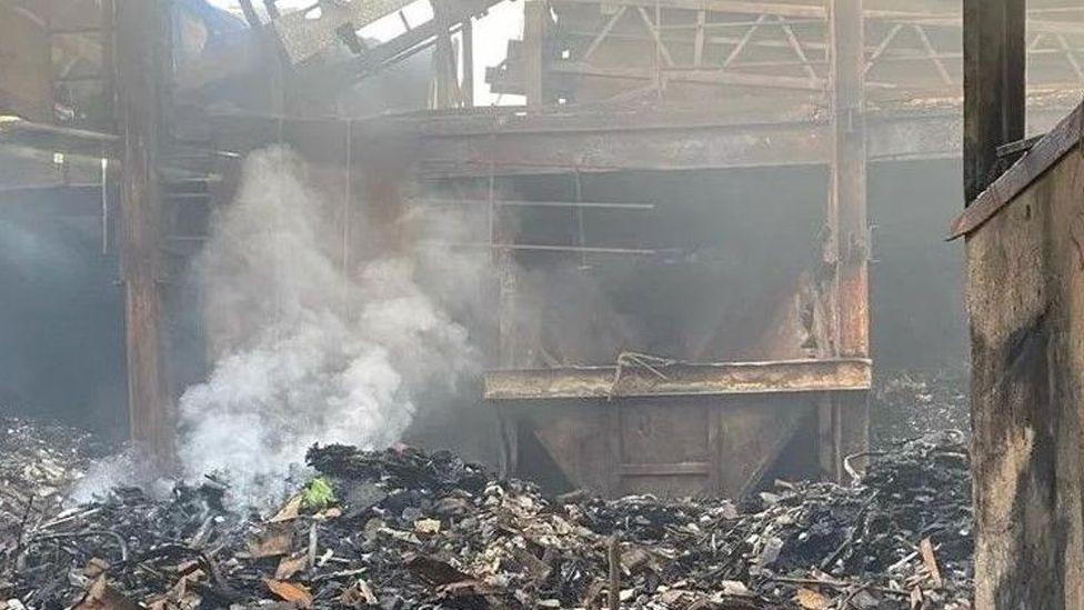 The aftermath of the fire showing smouldering waste, a burned out skip and the remnants of a building ravaged by fire