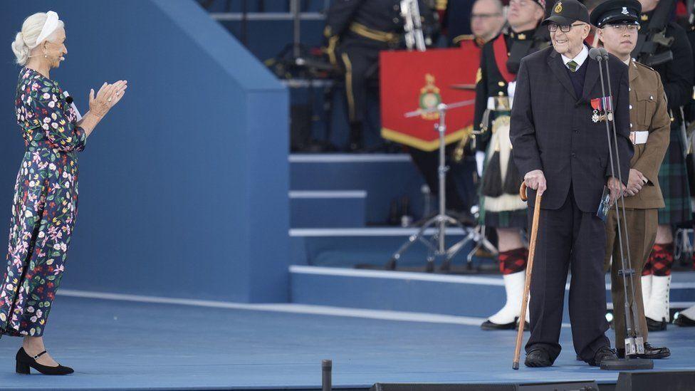 Stage with band members in background - man with walking stick to right wearing navy suit adorned with medals - Dame Helen Mirren in floral dress is clapping towards the man.