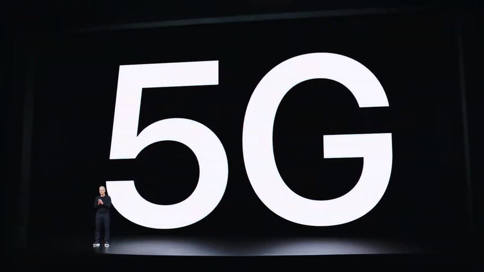 The letters 5G are displayed on screen behind Tim Cook on a huge dark empty stage