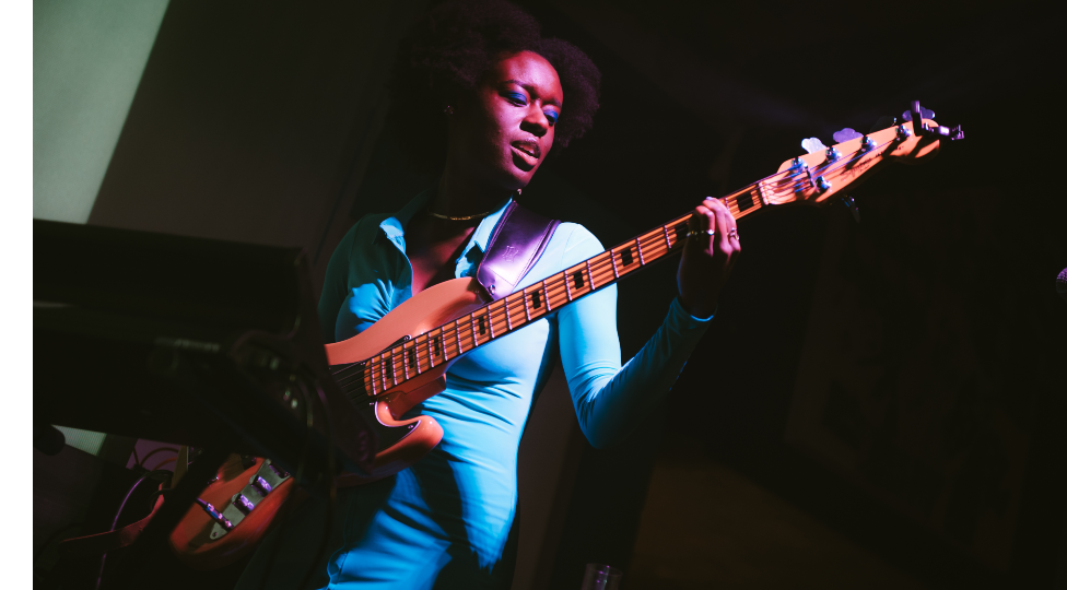 A woman in a blue jumpsuit is on a dark stage, playing the bass. Only her and her bass are lit up.