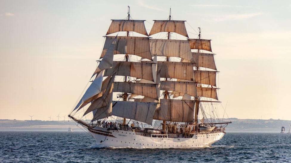 Tall cream-coloured ship - Sorlandet - with masts and beige sails at sea, with land and wind turbines in the distance.