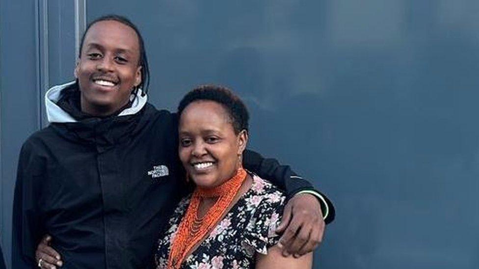 A young black man and a middle aged black woman with their arms around each other smiling