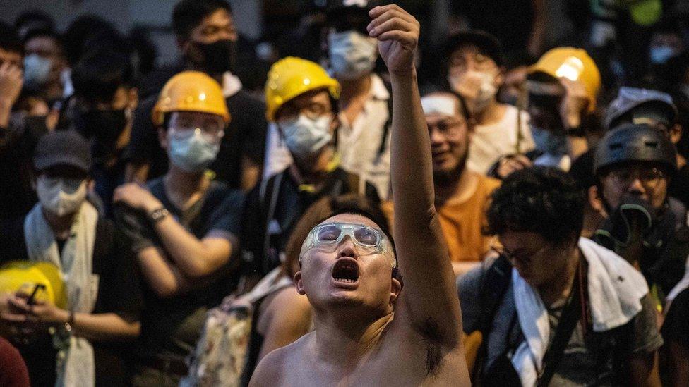 A shirtless man wearing swimming goggles raises one hand to the sky, eyes closed, moth open, while a crowd, many wearing yellow hard-hats, watch in the background