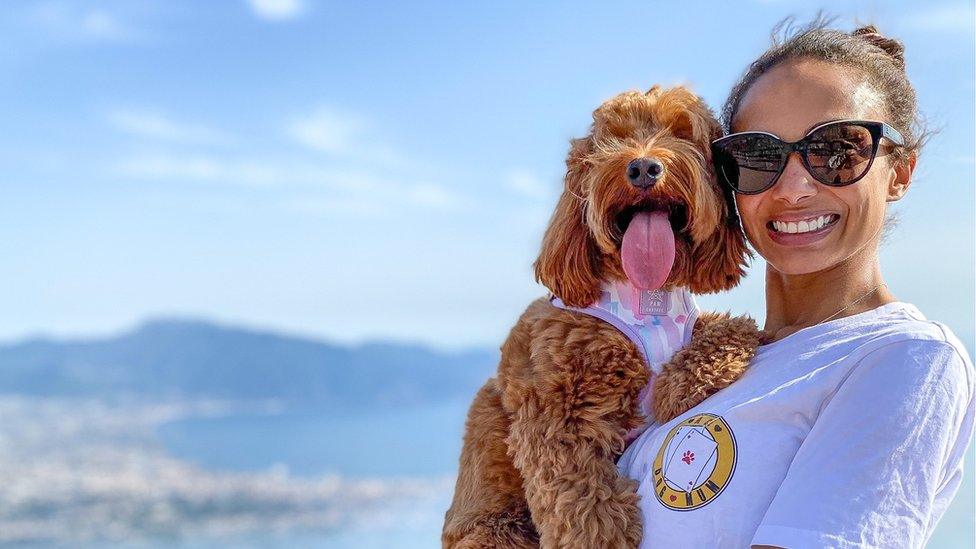 Mary McCall wears sunglasses and holds her red cockapoo Nola in her arms by Italian coastline