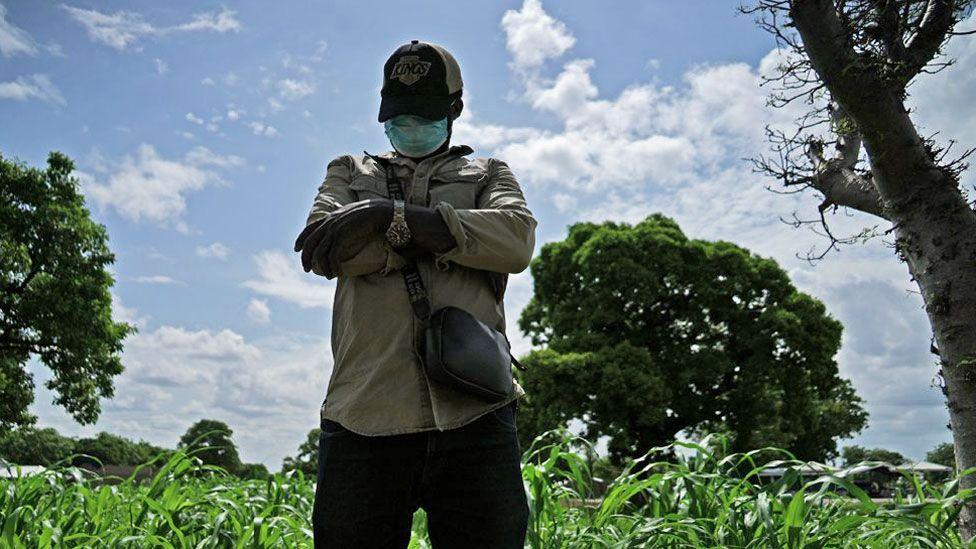 James (not his real name) looks down as he poses for a photo with his arms folded wearing a wrist watch, baseball cap, face mask, trousers, a khaki shirt and small crossbody brown bag. He is standing in a maize field with big trees and a blue sky in the background