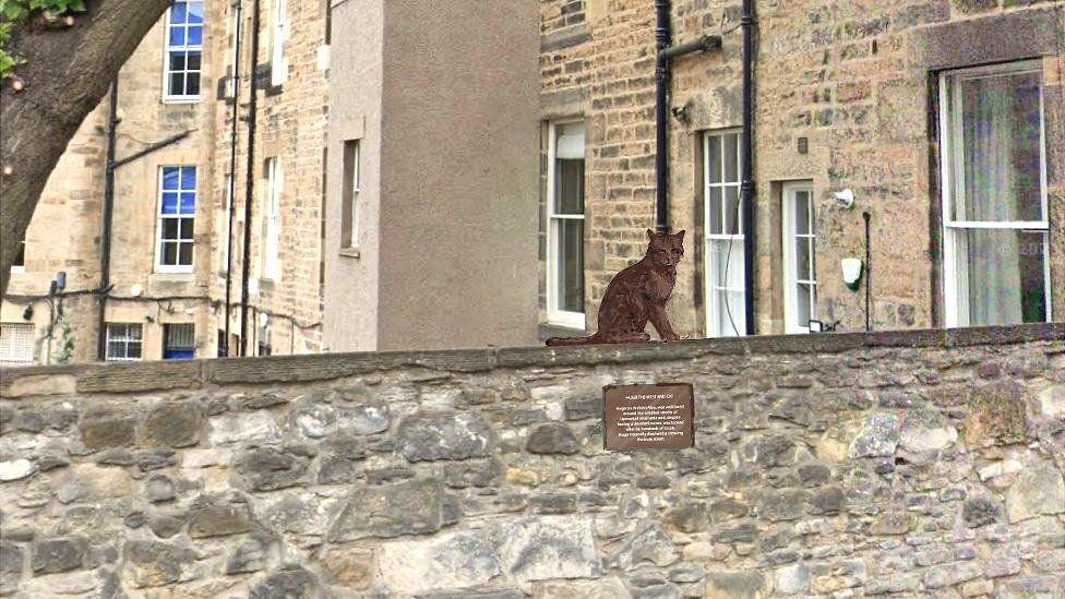 A drawing showing how the brown cat would be sitting on the wall. There is a plaque underneath but you can't make out the writing on it. There are tenement flats in the background.