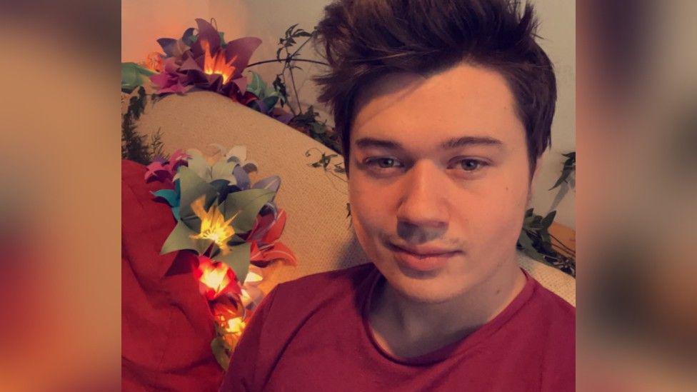 A selfie of a man with dark brown hair wearing a red T-shirt. He is sitting on a sofa with artificial light-up flowers on it.