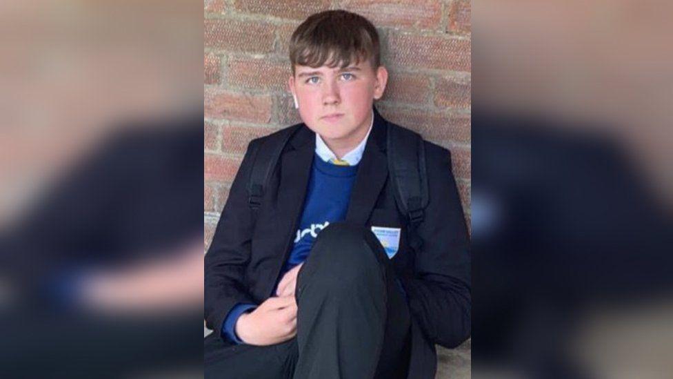 Harley Barfield sitting against a wall. He has short brown hair and is wearing a school uniform, including a blue jumper, with a black pack back still on his back. 