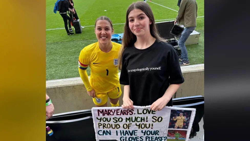 Mary Earps in a yellow England goalkeeper kit poses for a photo with Emmy, who is dressed in a black shirt with the words "be unapologetically yourself" written in white across the centre. Emmy is holding a homemade sign which says "Mary Earps, love you so much! Can I have your gloves please?