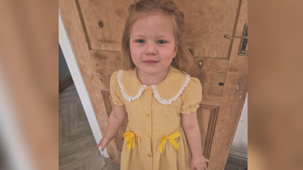 A five-year-old girl with long blonde hair wearing a yellow gingham dress with yellow bows on it and a frilly white collar. She is smiling and stood in front of a brown wooden door.