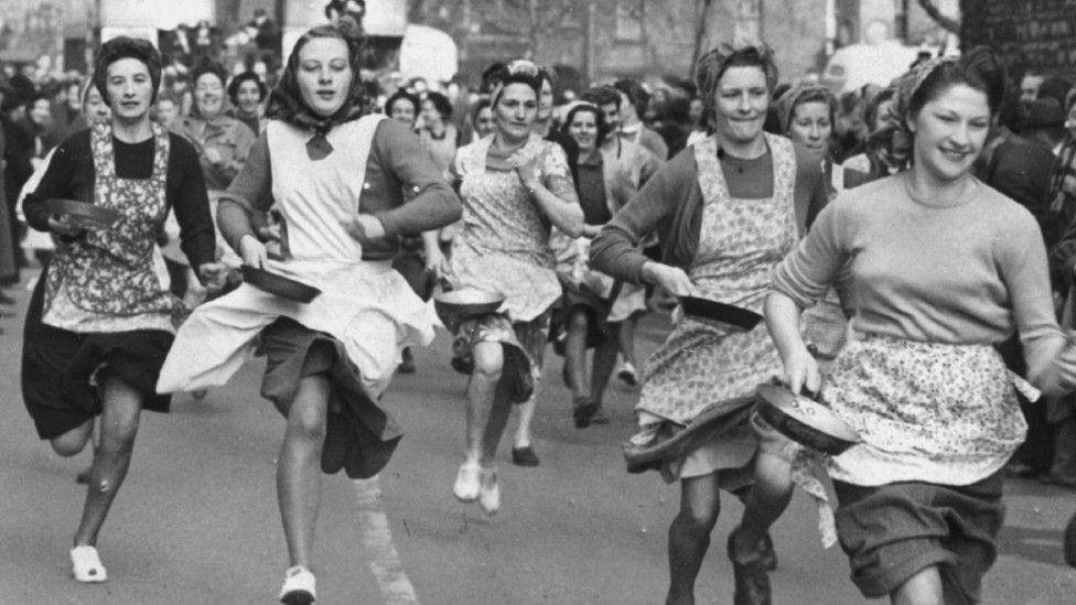 A group of women taking part in a pancake race. Rules of the tradition states the women must be wearing a skirt, apron and headscarf and must be carrying a pan with a pancake inside. There are too many women in the picture to count and it is black and white.