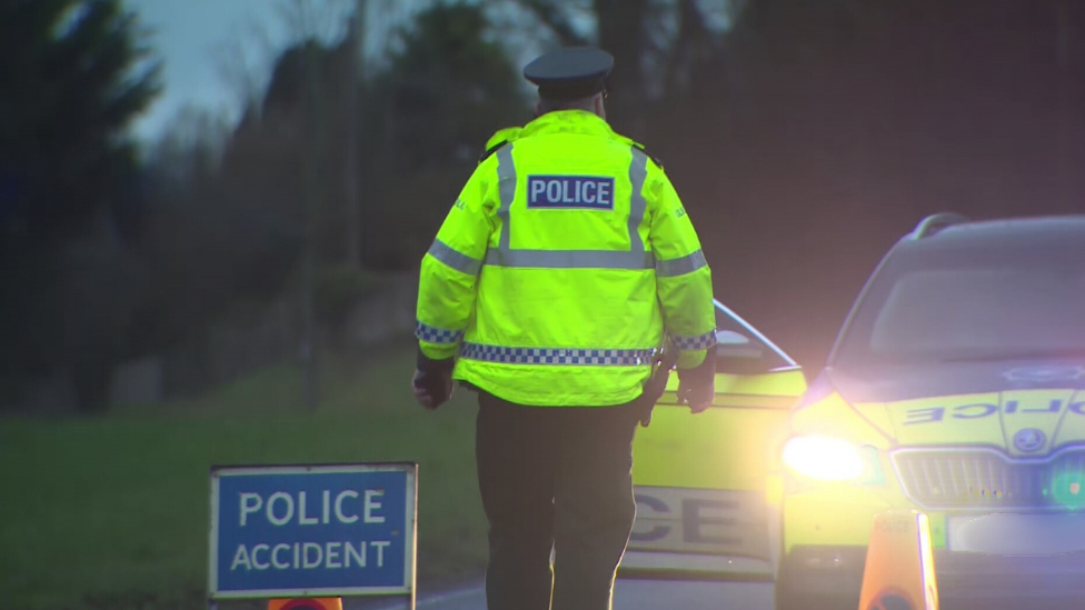 Police accident road sign, back of a policeman wearing a high viz police jacket and a police car visible behind him, yellow traffic cone showing road closure