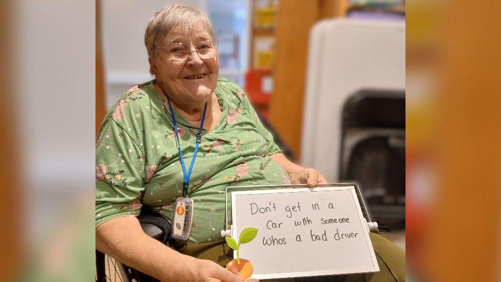 A woman with short grey hair and wearing a green top and glasses. She's sitting in a wheelchair in a room, and holding up a whiteboard saying "don't get in a car with someone who's a bad driver".