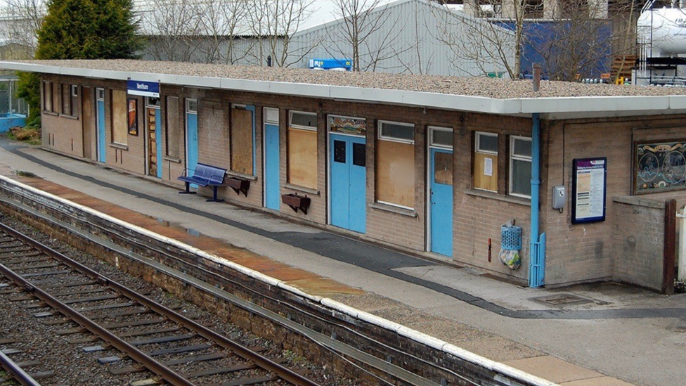 A railway station building with boarded-up windows and locked doors. There is graffiti on parts of the building, and the platform is uneven and discoloured. Planters attached to the wall are empty.