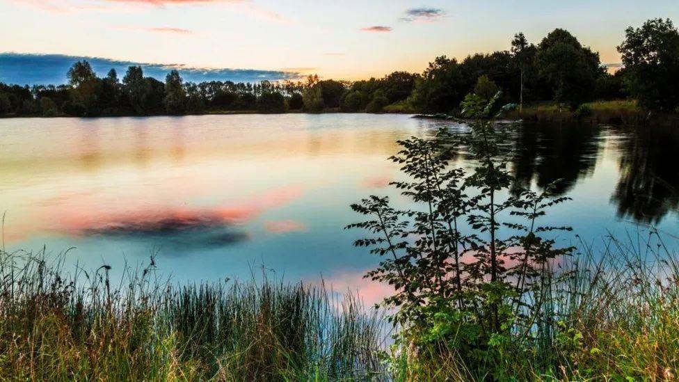 Cotswold Water Park. The sunset is reflecting in the water, casting shades of pink, orange and yellow. Trees are also reflecting in the water.