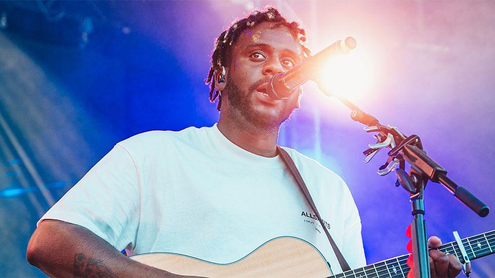 Myles Smith, wearing a light blue tshirt, singing on stage into a black microphone, while playing a guitar. There is a bright spotlight shining behind him.