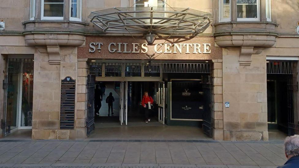 The front of the St Giles Centre - its name in bold letters - in Elgin, with a  woman in a red coat walking through the open front doors.