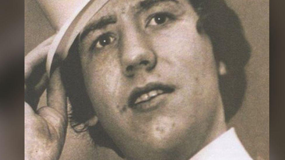 Columba McVeigh, looking to the right of camera, with his mouth slightly open.  He is wearing a white shirt and his hand is holding onto a had which is on his head.  