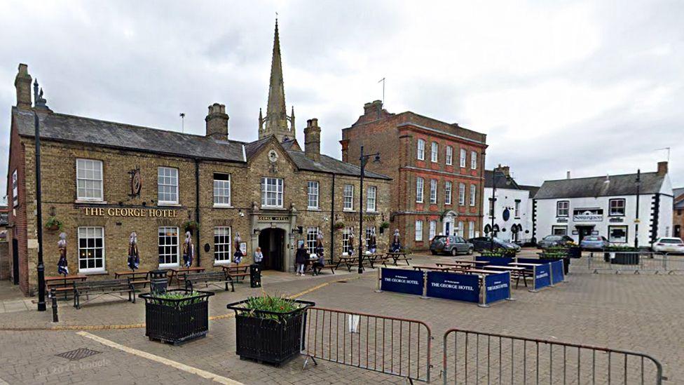 Whittlesey Market Place, showing the George Hotel and a church spire in the backdrop. There are people milling about and outdoor benches to eat on.