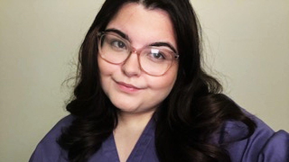 Ayla Jones is a white woman with long dark hair and large light brown framed glasses. She is wearing a purple medical scrubs top and is looking slightly to the right of the picture and smiling.