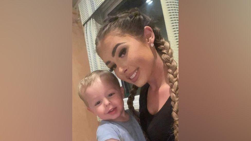 Grace Robinson is smiling at the camera in a selfie and is carrying a small boy in he arms. She has ling brown hair in plaits and the child has blonde hair and is wearing a blue T-shirt