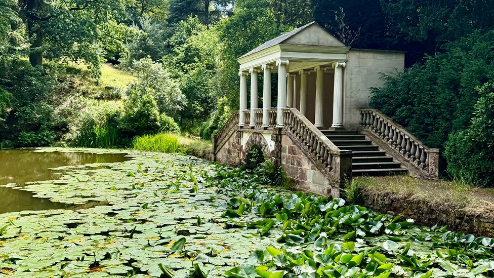 A secluded pool is covered in water lilies and other floating plants, with stone steps rising to a small, porticoed structure on the right designed in a classical style with white ionic columns supporting the roof. Woodland lies behind the building and fringing the bank of the pool on the left